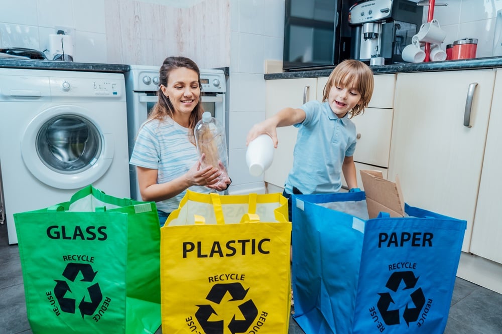 Mother is teaching kid how to recycle help the boy aware environmental importance - mom educates son sort garbage into different bins on kitchen. Family sorting garbage at home. Concept of recycling